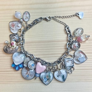 May include: A silver-toned charm bracelet with various charms, including heart-shaped pendants, oval pendants, and round pendants. The charms feature floral and scenic designs in pastel colors. The bracelet has a chain-link design and a lobster clasp.
