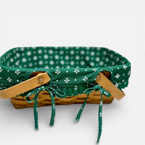 May include: A rectangular picnic basket with a woven brown exterior. The basket features wooden handles and is lined with a green fabric patterned with white floral designs. Green fabric ties secure the lining.