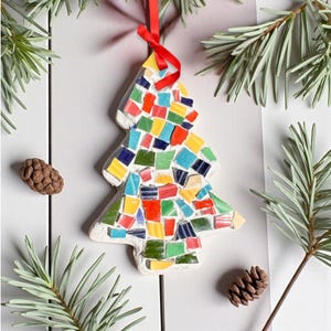 May include: A mosaic Christmas tree ornament with a red ribbon for hanging. The tree is made of colorful, irregularly shaped tiles in shades of red, blue, yellow, and green. The ornament is surrounded by pine needles and pine cones.