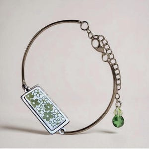 Op de afbeelding: Een zilverkleurige metalen armband met een rechthoekige bedel met een witte achtergrond en een herhalend patroon van olijfgroene bloemen. De armband bevat een kettingverlenging met een kleine, gefacetteerde lichtgroene en donkergroene kraal.