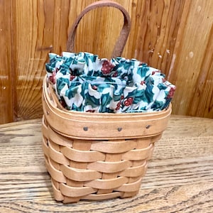 May include: A small, woven basket with a brown leather handle. The basket's interior is lined with a fabric featuring a white floral pattern with green leaves and red berries. The basket is made of light brown wood.