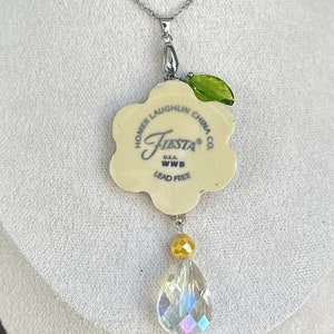 Puede incluir: Un colgante de cerámica blanca con forma de flor con el texto "Homer Laughlin China Co. Fiesta® U.S.A. WWB Lead Free" en la parte posterior. El colgante está unido a una cadena plateada con una hoja de vidrio verde y un colgante de cristal transparente en forma de lágrima.