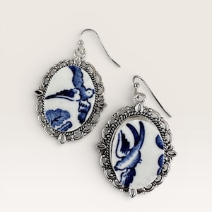 May include: A pair of silver-toned drop earrings. Each earring features an oval porcelain insert with a blue and white bird design. The porcelain is set in a decorative silver frame. The earrings have a hook closure.
