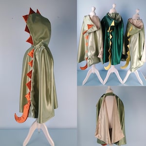 May include: Olive green dinosaur cape with orange detailing, including a spine and tail. The cape has a hood and is displayed on a white mannequin. Other capes in green and dark green are also shown.