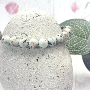 May include: A beaded bracelet with round, light blue and white beads with black speckles. The bracelet is draped over a textured, light gray stone. Green leaves and a soft, blurred background complete the image.