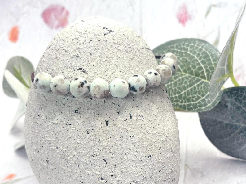 May include: A beaded bracelet with round, light blue and white beads with black speckles. The bracelet is draped over a textured, light gray stone. Green leaves and a soft, blurred background complete the image.