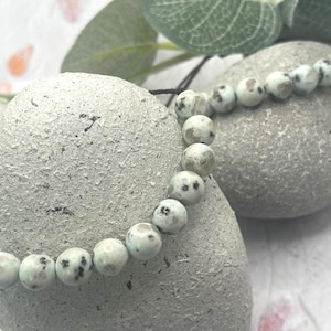 May include: A beaded bracelet featuring round, light-colored beads with dark spots. The bracelet is strung on a dark cord and rests on a textured, gray stone. Green leaves are visible in the background. The bracelet is a jewelry item.