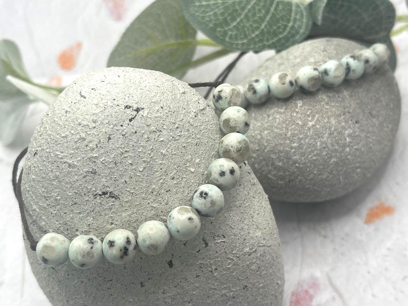 May include: A beaded bracelet featuring round, light-colored beads with dark spots. The bracelet is strung on a dark cord and rests on a textured, gray stone. Green leaves are visible in the background. The bracelet is a jewelry item.