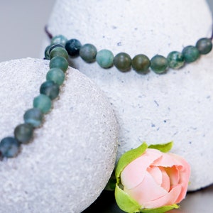 May include: A green agate beaded necklace with a brown cord on a white textured surface. A pink rose bud is in the foreground.