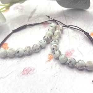 May include: Two beaded bracelets with light blue and gray speckled stones. The beads are strung on a dark brown cord with adjustable sliding knots. The bracelets are displayed on a textured white surface.