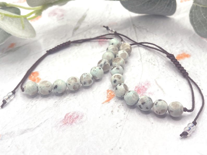 May include: Two beaded bracelets with light blue and gray speckled stones. The beads are strung on a dark brown cord with adjustable sliding knots. The bracelets are displayed on a textured white surface.
