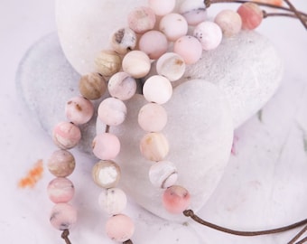 May include: A strand of pink and white opal beads on a brown leather cord. The beads are arranged in a graduated pattern, with the larger beads at the top and the smaller beads at the bottom. The beads are resting on a white surface.