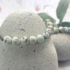 May include: A beaded bracelet featuring round, light green beads with dark gray speckles. The bracelet is strung on a brown cord and rests on a textured, gray stone. The image has a soft, natural aesthetic.