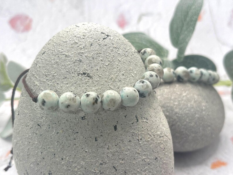 May include: A beaded bracelet featuring round, light green beads with dark gray speckles. The bracelet is strung on a brown cord and rests on a textured, gray stone. The image has a soft, natural aesthetic.