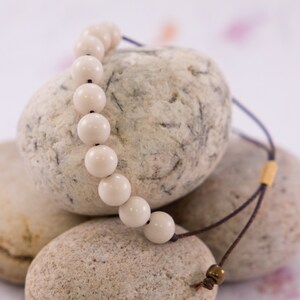 May include: A white beaded necklace with a brown cord and gold accents. The necklace is draped over three smooth, light brown stones.
