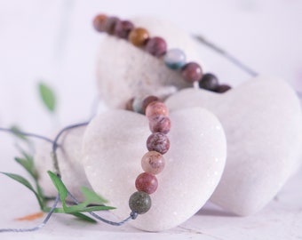 May include: A heart-shaped white stone with a multi-colored beaded necklace draped over it. The necklace is made of small, round beads in shades of brown, red, and green.