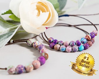 May include: Two bracelets with purple, blue, and brown jasper beads on brown cords. The bracelets are on a white surface with a white flower and a gold star sticker that says "#1 Best Seller 10 Breaths Bracelet".