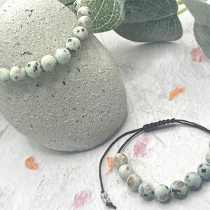 May include: Two beaded bracelets with a natural stone appearance. The beads are light blue with dark gray spots. One bracelet is draped over a textured gray stone, and the other lies flat. The bracelets are on a white surface with floral accents.