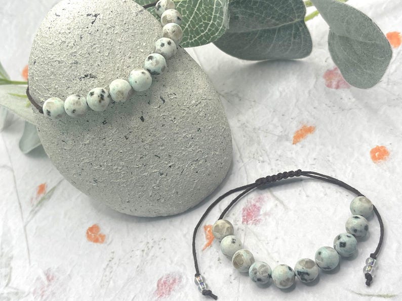 May include: Two beaded bracelets with a natural stone appearance. The beads are light blue with dark gray spots. One bracelet is draped over a textured gray stone, and the other lies flat. The bracelets are on a white surface with floral accents.