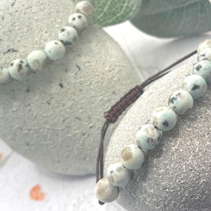 May include: Two beaded bracelets with light blue and brown speckled beads. The bracelets are on a gray stone surface. One bracelet has a brown adjustable cord. The beads have a diameter of approximately 0.3 inches.