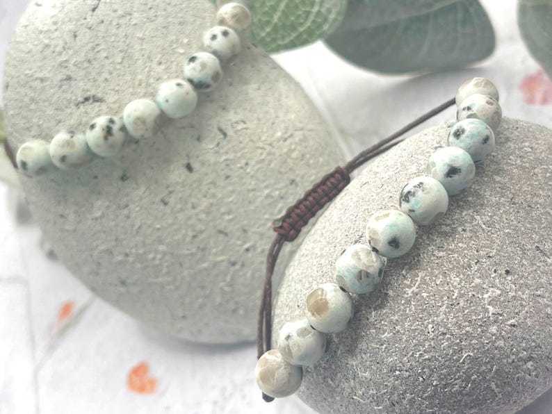 May include: Two beaded bracelets with light blue and brown speckled beads. The bracelets are on a gray stone surface. One bracelet has a brown adjustable cord. The beads have a diameter of approximately 0.3 inches.