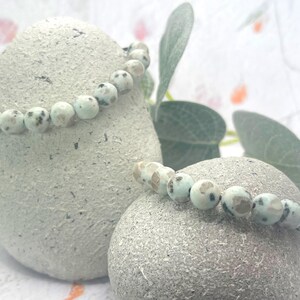 May include: Two beaded bracelets with light blue and brown speckled beads are displayed on textured, light gray stones. The bracelets are strung on a black cord. Green leaves and a soft, blurred background complete the image.