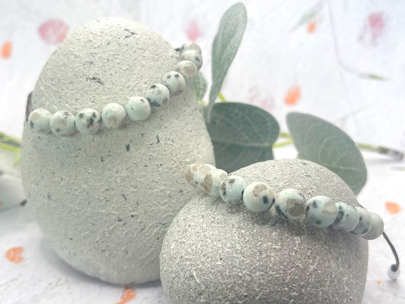 May include: Two beaded bracelets with light blue and brown speckled beads are displayed on textured, light gray stones. The bracelets are strung on a black cord. Green leaves and a soft, blurred background complete the image.