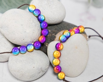 May include: Two strands of iridescent rainbow beads on a brown cord. The beads are round and have a metallic finish. The beads are arranged in a gradient pattern, starting with yellow and transitioning to purple.