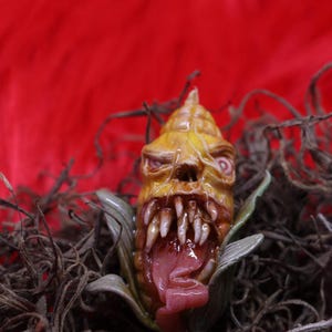 May include: A grotesque, yellow corn sculpture with sharp teeth and a protruding pink tongue. The corn is set against a red background and surrounded by dried plant matter, creating a spooky, Halloween-themed aesthetic.