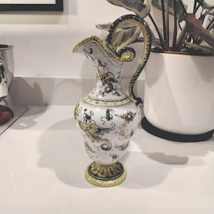 Puede incluir: Una jarra de cerámica blanca con un diseño floral amarillo y marrón. La jarra tiene un asa curva y una base ancha.