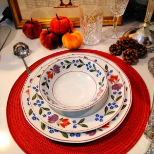 以下が含まれることがあります： 青、赤、緑の花柄が描かれた白いセラミック製のプレート3枚セット。プレートは重ねて置かれ、赤いランチョンマットの上に置かれています。