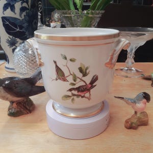 Może przedstawiać: Biała ceramiczna donica ze złotymi obramowaniami i uchwytami, z namalowanym wzorem ptaków na gałęzi. Donica stoi na białym obrotowym stojaku, w pobliżu inne dekoracyjne figurki ptaków.