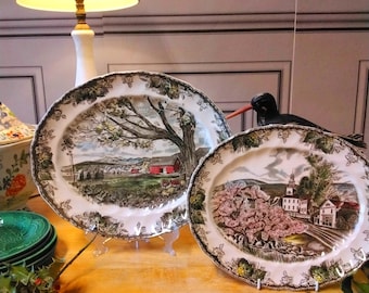Pode incluir: Dois pratos de servir ovais de cerâmica com um design de cena campestre. O prato à esquerda apresenta um celeiro vermelho e uma árvore em um campo verde. O prato à direita apresenta um edifício branco com uma árvore rosa em primeiro plano.