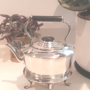 May include: A silver teapot with a black handle and a black knob on the lid. The teapot is sitting on a silver stand with three legs.