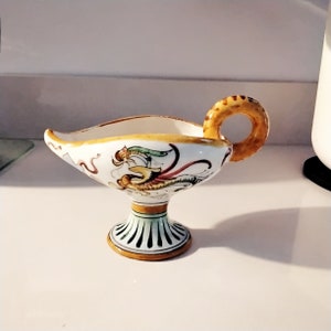 Puede incluir: Una salsera de cerámica blanca con un diseño floral amarillo, verde y rojo. La salsera tiene un asa en forma de anillo y una base de pedestal.