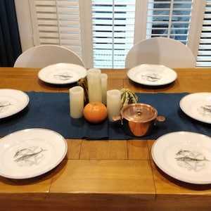 Puede incluir: Una mesa de madera preparada para una cena con sillas blancas, manteles individuales azules, platos blancos con diseño de pescado y platos de servir de cobre. También hay velas y calabazas en la mesa.