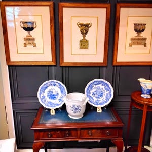 May include: Three framed prints of antique urns with gold accents, displayed on a wall with dark blue wainscoting. Below the prints, a wooden table with two blue and white porcelain plates and a white porcelain pitcher with blue floral designs.