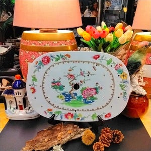 May include: An oval, white ceramic serving platter with a floral design in pink, red, yellow, and green. The platter has a brown border and small holes around the edge. It is displayed on a black stand.