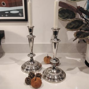 May include: Two silver candlesticks with white candles, featuring a beaded design on the base and top. The candlesticks are displayed with small pumpkins and pinecones, creating a fall-themed arrangement.
