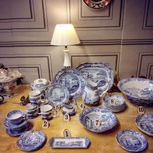 Op de afbeelding: Een verzameling blauw-wit gepatroneerd keramiek, waaronder een grote schotel, een kan, kommen, mokken en schoteltjes. Het serviesgoed heeft een landschapsontwerp met bomen, een brug en een gebouw. Het serviesgoed is gerangschikt op een houten tafel met een lamp op de achtergrond.
