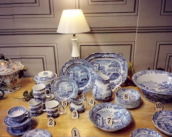 May include: A collection of blue and white patterned ceramic dishes, including a large platter, a pitcher, bowls, mugs, and saucers. The dishes feature a scenic landscape design with trees, a bridge, and a building. The dishes are arranged on a wooden table with a lamp in the background.