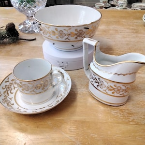 May include: A white teacup and saucer with a gold floral design, a white creamer with a gold floral design, and a white sugar bowl with a gold floral design.