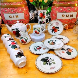 Puede incluir: Un conjunto de vajilla de cerámica blanca con diseño de cerezas. El juego incluye una tetera, tazas, platillos, una jarra de crema y tazas. En el fondo, barriles decorativos con las palabras "GIN" y "RUM".