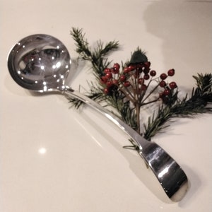 May include: A polished silver ladle with a circular bowl and a slender handle. The ladle rests on a white surface, accompanied by a sprig of evergreen and red berries. This serving utensil is suitable for soups or sauces.