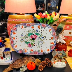 May include: An oval, white decorative table top with a floral design in pink, blue, and green. The table top has a series of small holes and is surrounded by a floral border. The table top is displayed with other decorative items, including lamps and figurines.