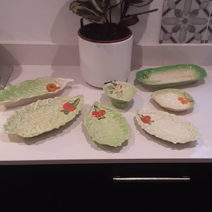 Può includere: Una collezione di piatti da portata in ceramica a forma di foglie di lattuga, con un accento di pomodoro. I piatti sono in varie tonalità di verde e bianco. Una pianta in vaso è sullo sfondo.