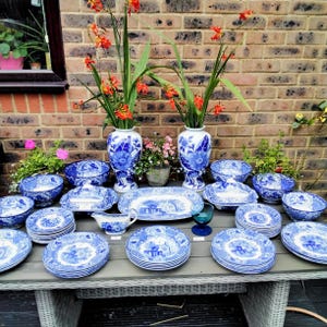 Puede incluir: Un conjunto de vajilla de porcelana azul y blanca, que incluye platos, cuencos y fuentes. Los platos presentan un diseño detallado de paisaje. Dos jarrones con flores naranjas se exhiben sobre una mesa gris.