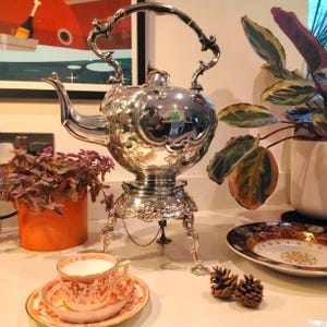 May include: A silver teapot with ornate details and a three-legged stand. The teapot is polished and reflects the surrounding environment. The teapot is on a white surface with a teacup and saucer, pine cones, and a decorative plate.