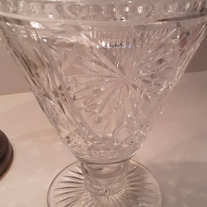 Puede incluir: Un cuenco de cristal tallado transparente, con una parte superior ancha y acampanada y una base de pedestal. El cuenco presenta intrincados patrones de estrella y geométricos, creando un efecto brillante. La base tiene un diseño circular con ranuras verticales.