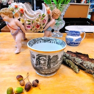 Könnte beinhalten: Ein dekorativer Keramik-Blumentopf mit einem schwarz-weißen Landschaftsdesign, akzentuiert mit einem goldenen Rand. Eicheln und ein kleiner blau-weißer Krug sind ebenfalls zu sehen. Ein von Engeln getragenes Blumengesteck ist ebenfalls sichtbar.